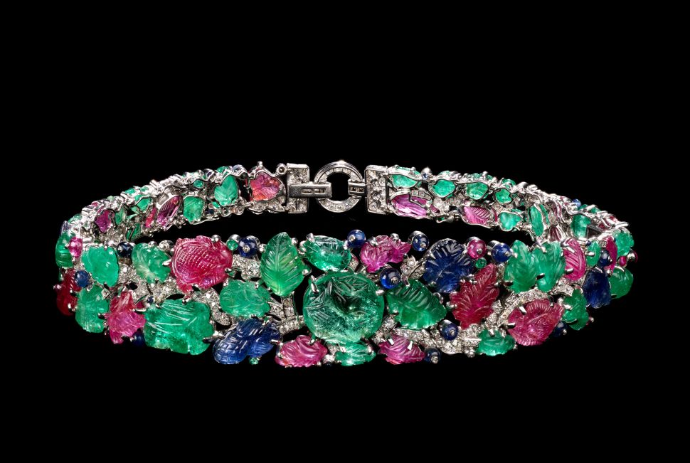 A close-up image of a jeweled Cartier bracelet from 1928 in the tutti-frutti style, made of carved emeralds, rubies, sapphires and diamonds set in platinum, displayed against a black background.