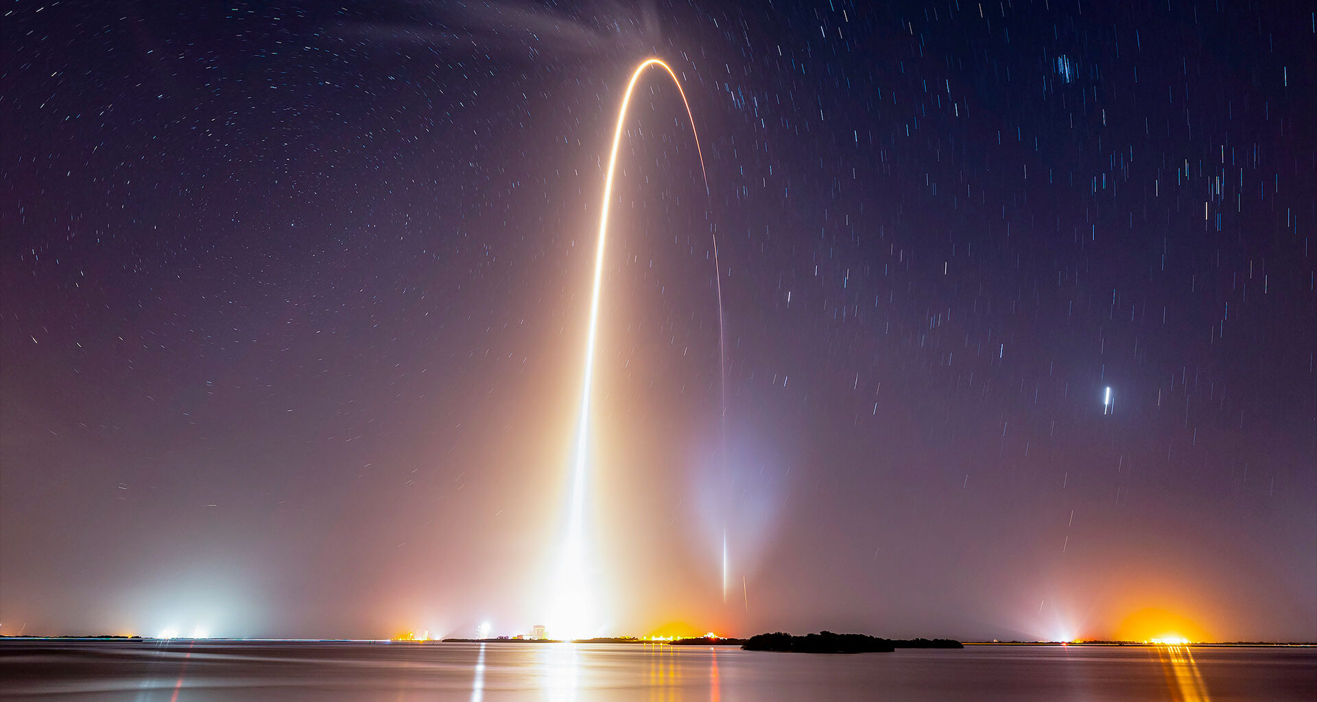 a time lapse view of a night time launch showing the arc of the rocket reflecting in body of water in the foreground