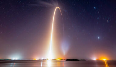 a time lapse view of a night time launch showing the arc of the rocket reflecting in body of water in the foreground