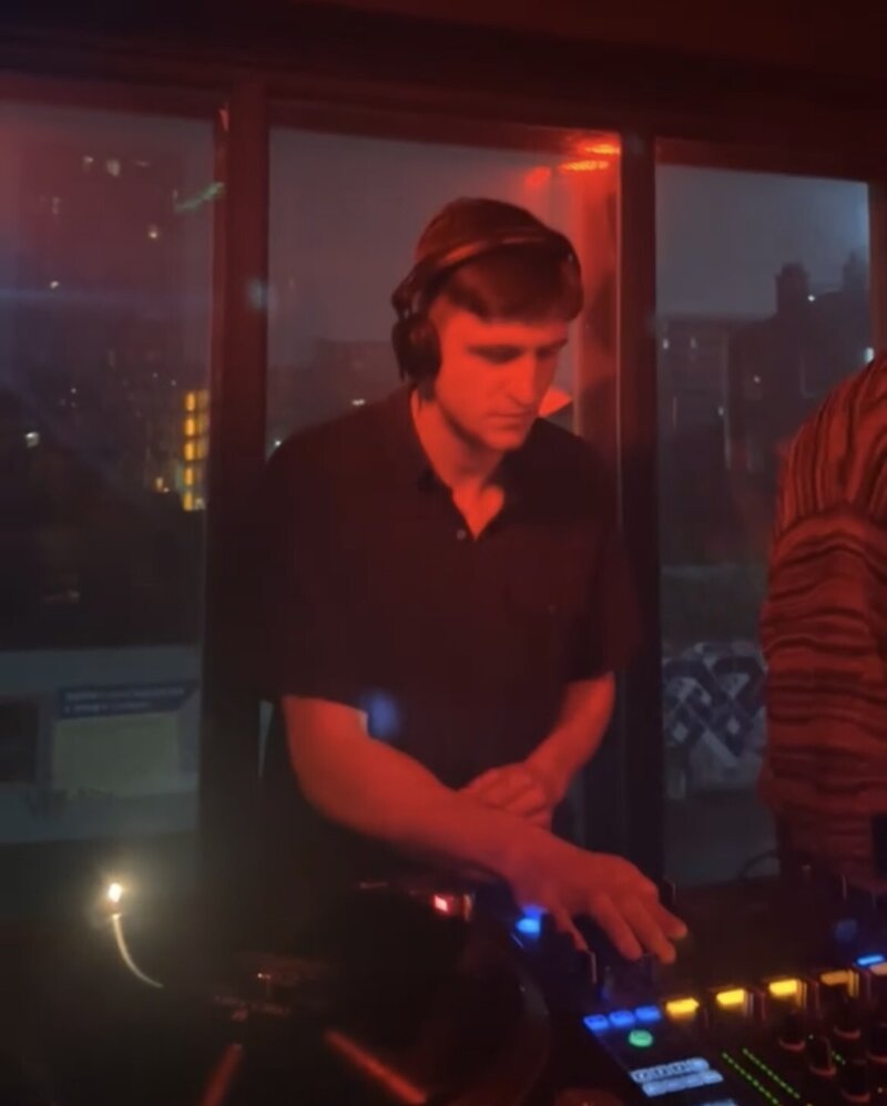 A close-up photo of a man DJing in a dark club, lit by red lights