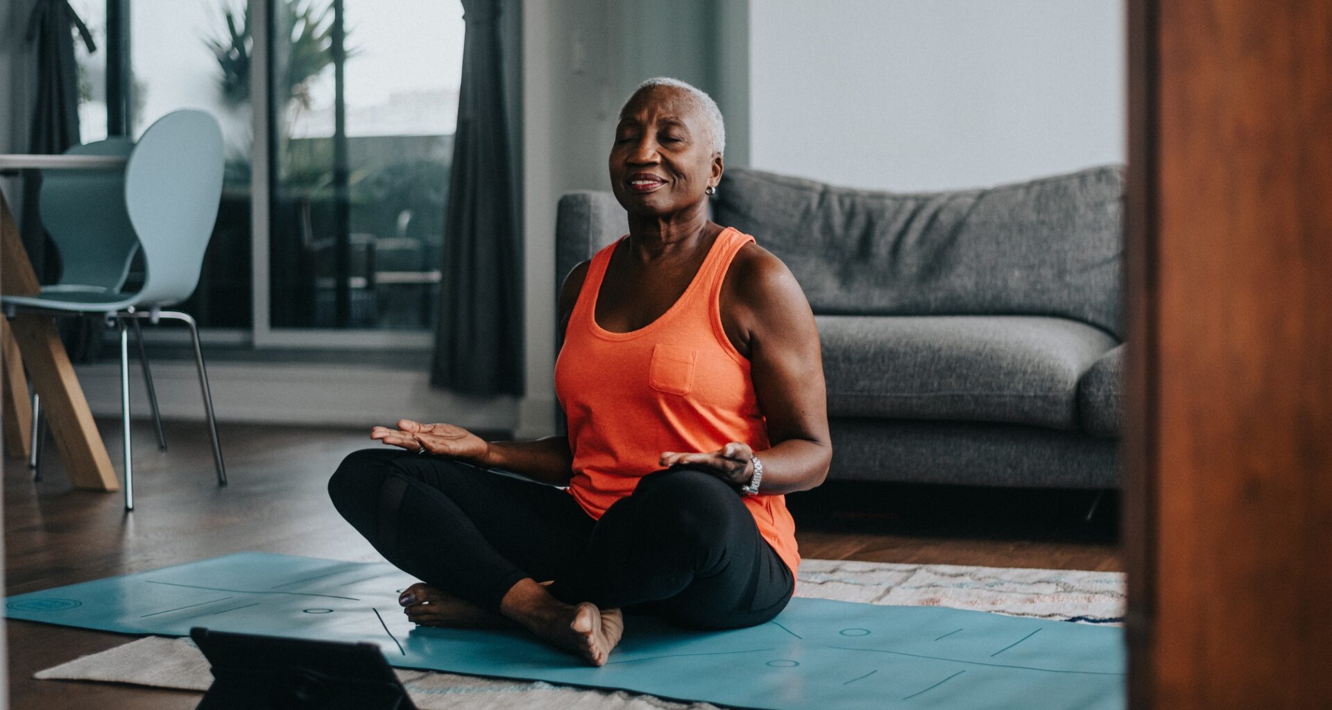 woman in a seated meditative posture on a blue mat in a living room with an ipad device in front of her