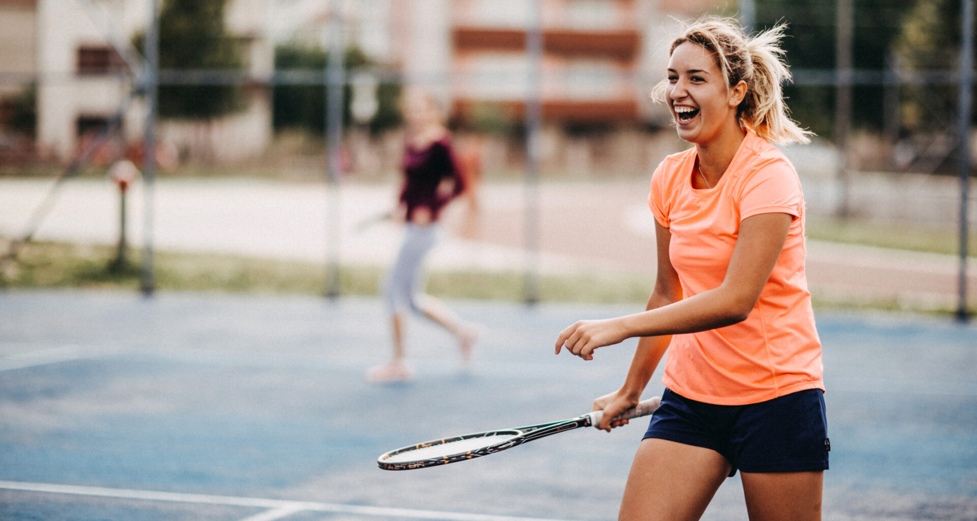 Woman holding tennis racket walking on tennis court. Her mouth is open and she is smiling