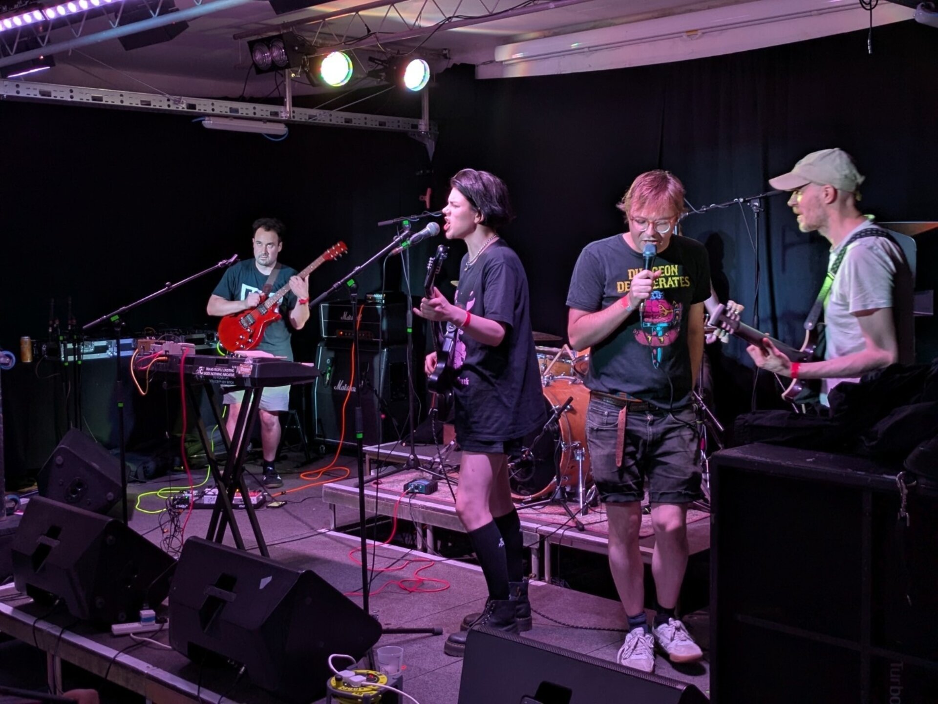 A photo of four people in a band playing onstage, including three guitarists or bassists and a vocalist, in a small music venue