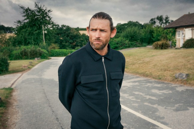 Chris Coghill playing mysterious new character, Kev, stands on the outskirts of the village, glancing sideways.
