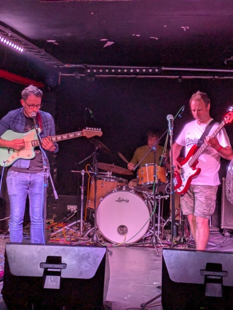 A close-up photo of a three-piece band performing onstage including a drummer, a bassist and a guitarist