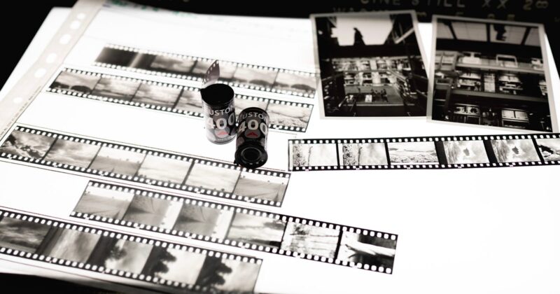Black and white photo negatives, film canisters, and two printed photographs are displayed on a lightbox, showcasing various urban and landscape scenes.