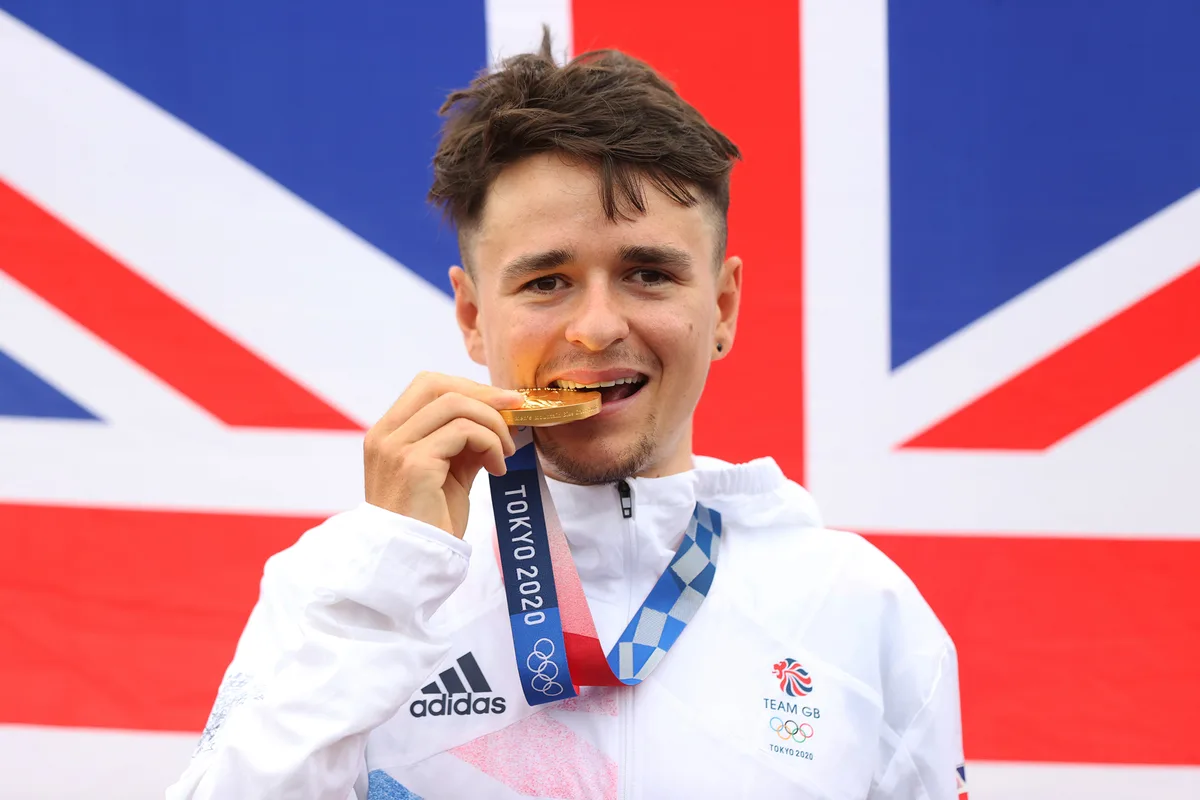 Thomas Pidcock of Team Great Britain bite his gold medal and pose with the flag of his country in the background after the Men's Cross-country race on day three of the Tokyo 2020 Olympic Games.