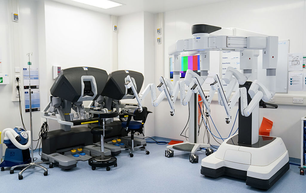 Image of a room in a hospital featuring two surgical robots and two control systems.