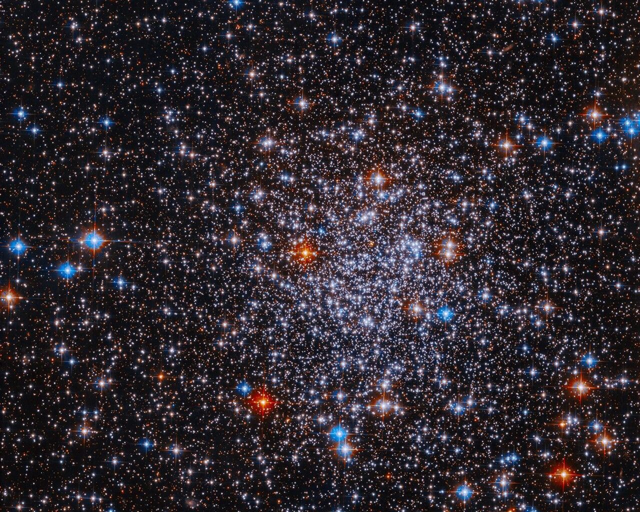 Bright stars cluster against a black background. The stars are more densely concentrated in the center of the image. The stars appear mostly white, but bright red and blue stars are also visible sprinkled throughout the image.