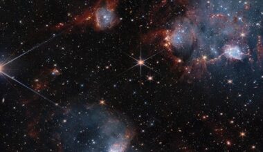 Bluish clouds of gas are wound through with red-glowing lanes and filaments of dust.. The clouds contain cavities filled with stars and many more stars are visible in the background and scattered throughout the clouds. The clouds are concentrated in the upper right and the lower middle of the image. Tiny background galaxies are also sprinkled throughout the image.