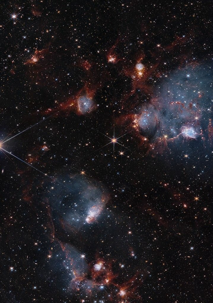 Bluish clouds of gas are wound through with red-glowing lanes and filaments of dust.. The clouds contain cavities filled with stars and many more stars are visible in the background and scattered throughout the clouds. The clouds are concentrated in the upper right and the lower middle of the image. Tiny background galaxies are also sprinkled throughout the image.