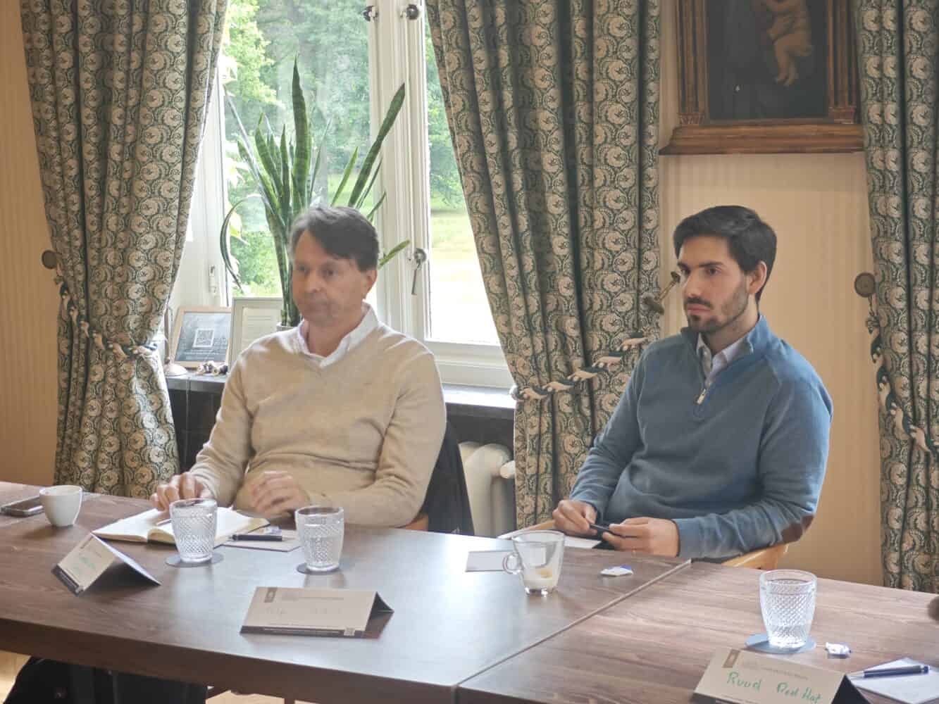 Twee mannen zitten aan een vergadertafel met papieren, glazen en naamplaatjes voor zich, in een kamer met gedessineerde gordijnen en een raam met groen buiten.