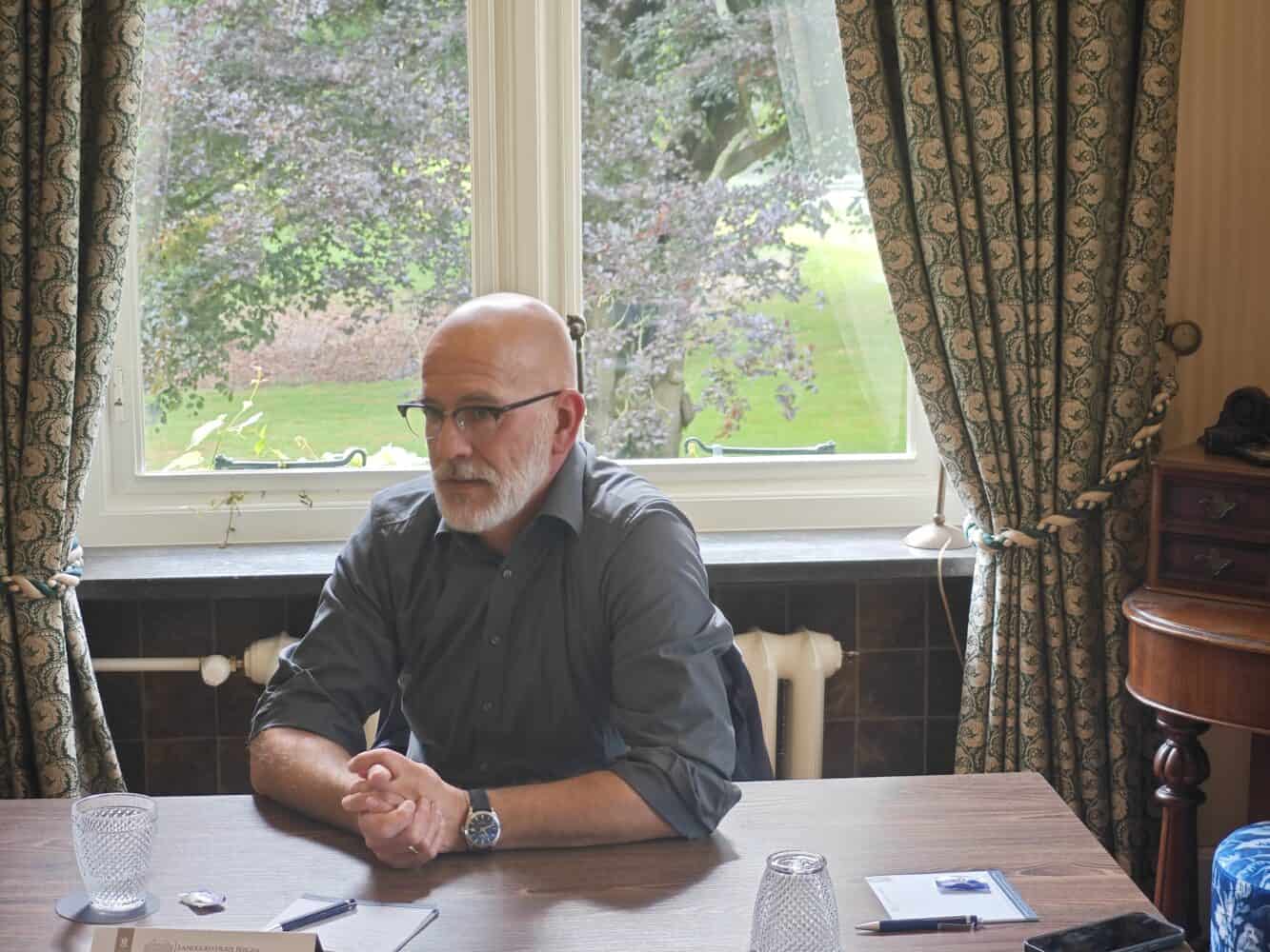 Een man met een bril en een grijs overhemd zit aan een houten tafel met papieren en glazen voor zich, in een kamer met grote ramen en gordijnen die bomen buiten laten zien.