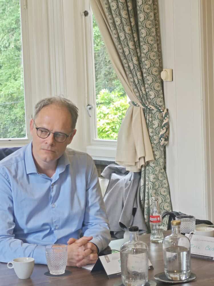 Een man in een blauw shirt zit aan een tafel met drinkglazen, waterflessen en een Coca-Cola fles voor een raam met gedessineerde gordijnen.