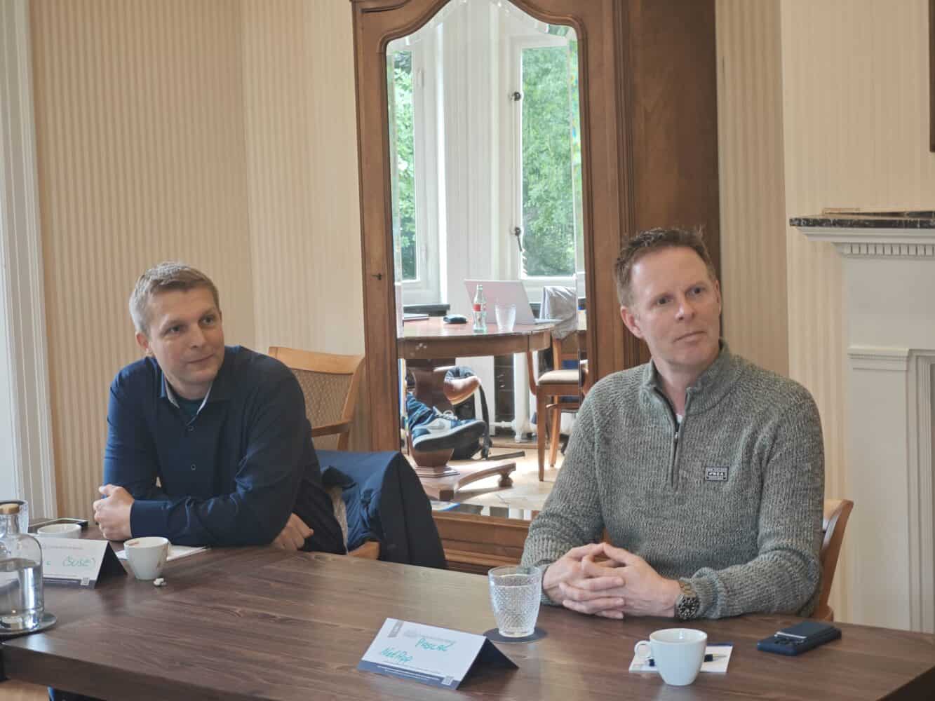 Twee mannen zitten aan een houten tafel met naamkaartjes, kopjes en glazen voor zich in een goed verlichte kamer met grote ramen en een spiegel.