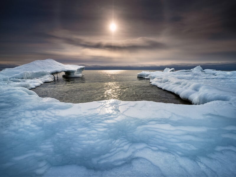 The image shows a frozen landscape with icy formations surrounding a pool of water. The sun is low in the cloudy sky, reflecting on the water and casting a serene, cold light across the icy scene.