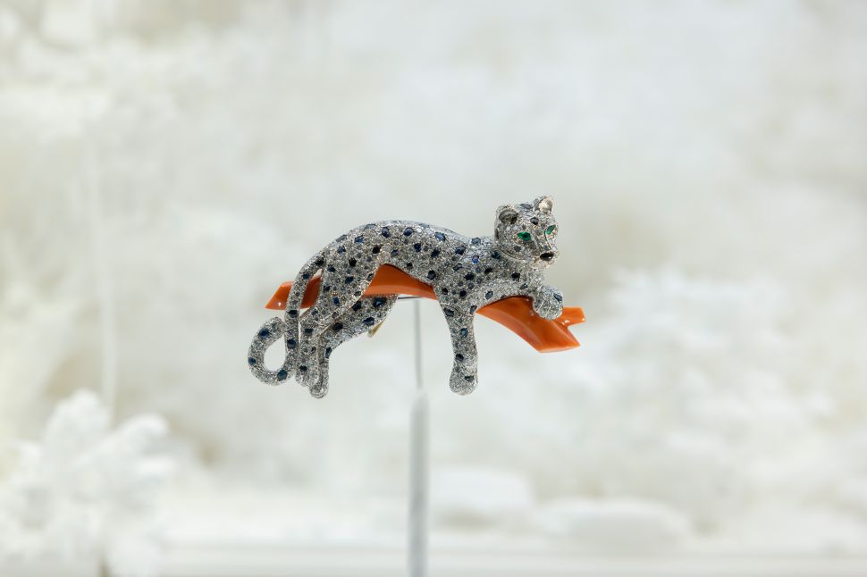 A Cartier brooch in the shape of a reclining panther made of diamonds and sapphires with emerald eyes is perched on a curved orange surface against a white background.