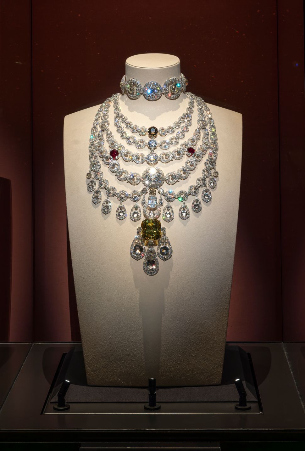An ornate Cartier necklace featuring large diamond loops and a prominent yellow central gem, known as the Palatia Necklace, is displayed on a white bust against a dark red wall.