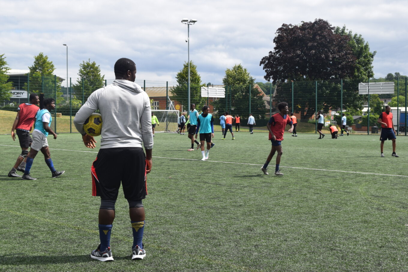 A man in a grey jumper looks on as others play a game of football, back to the camera and a football under his left arm.