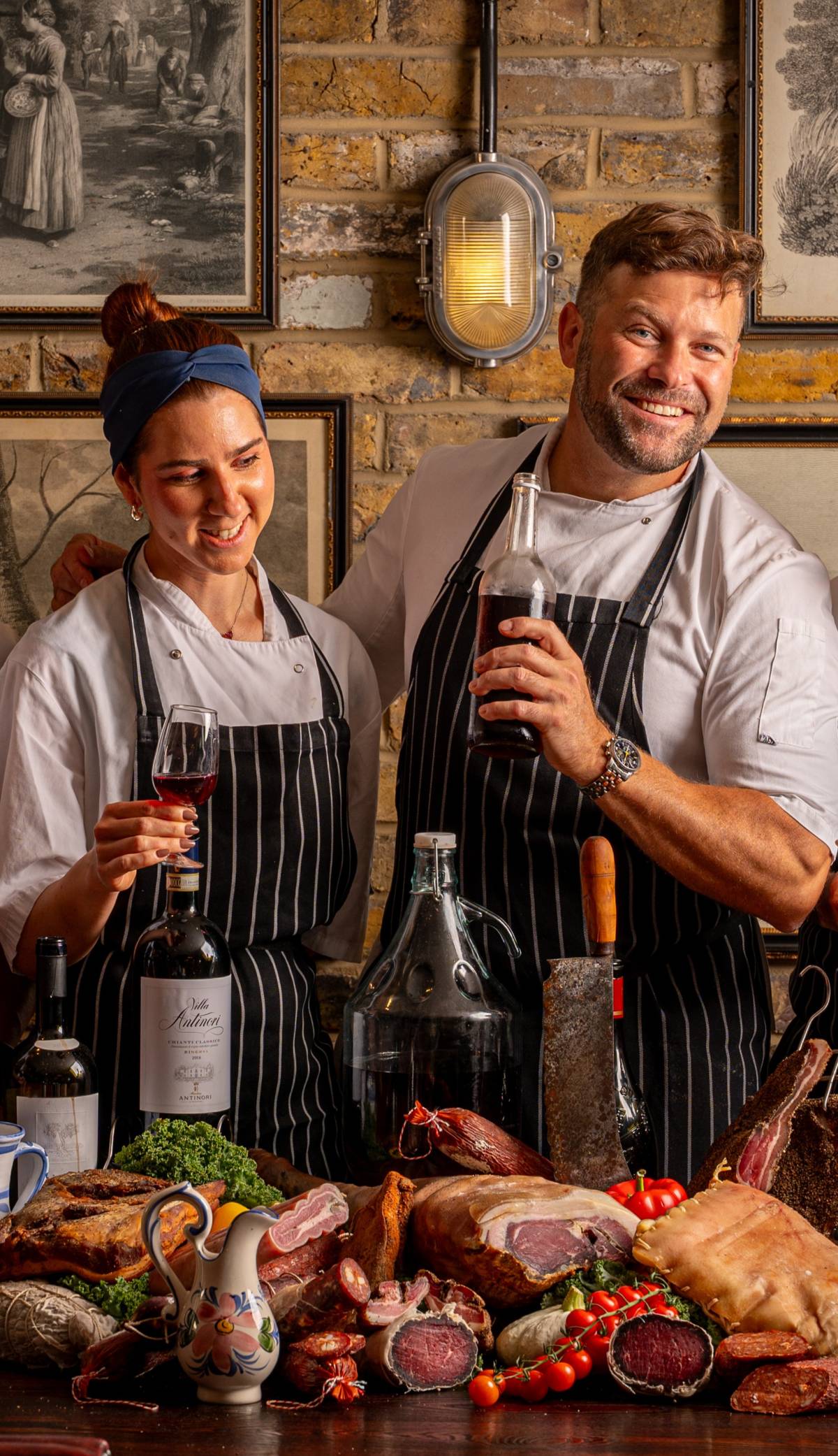 two chefs smile warmly, in front of a brick-walled background. the foreground shows glass bottles of varied shapes and sizes. they're smiling, while both holding a glass or bottle (each; respectively).