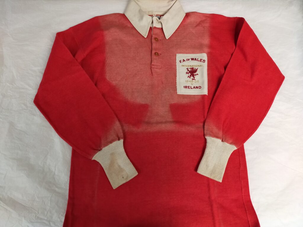 A vintage long-sleeve red rugby jersey with cream collar and cuffs, and a front pocket featuring the Welsh Rugby Union crest and the words Wales International Ireland. The shirt shows signs of fading and wear.