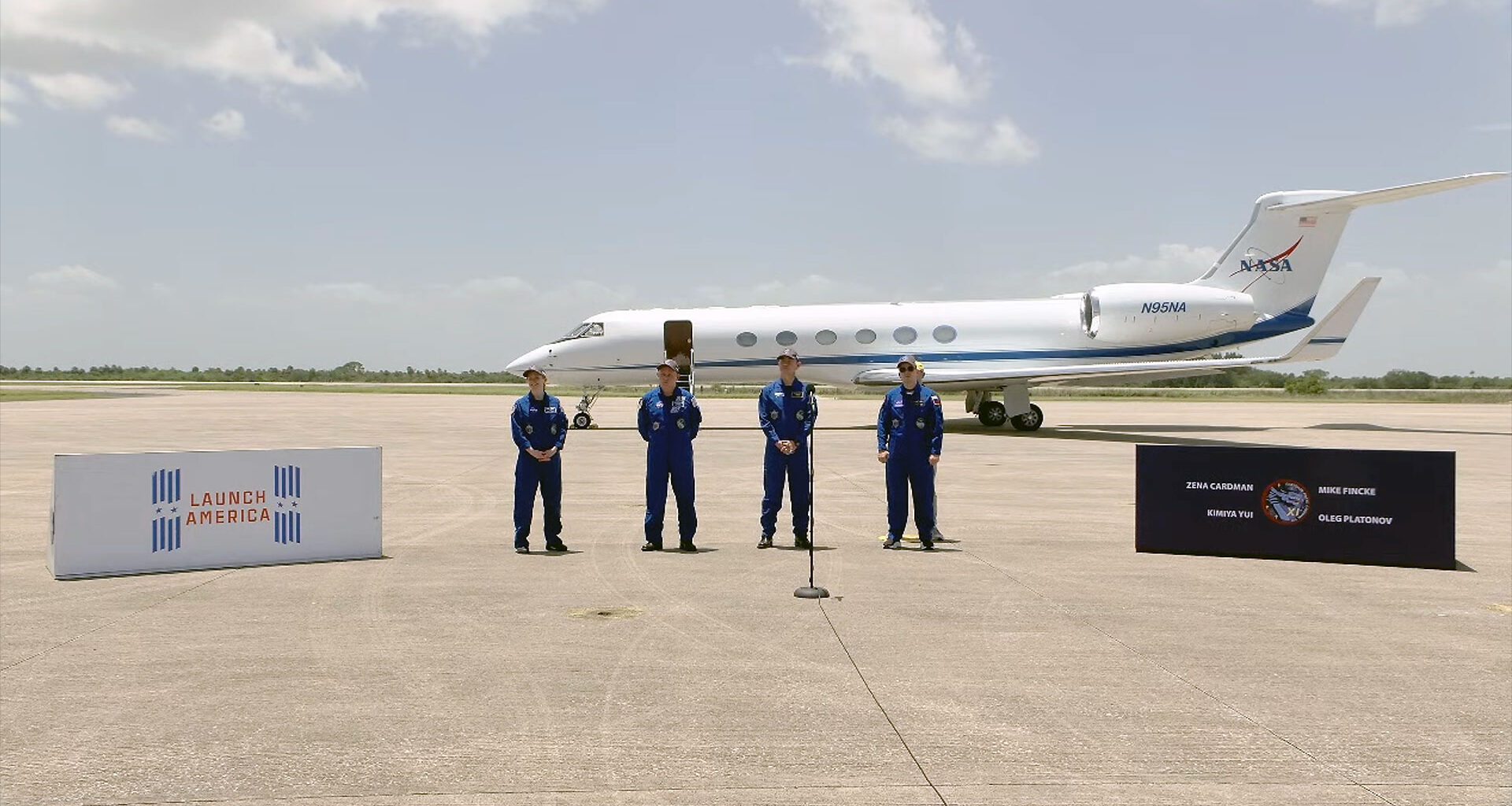 four people in blue flight suits stand on a tarmac in front of a small jet aircraft