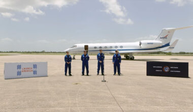 four people in blue flight suits stand on a tarmac in front of a small jet aircraft