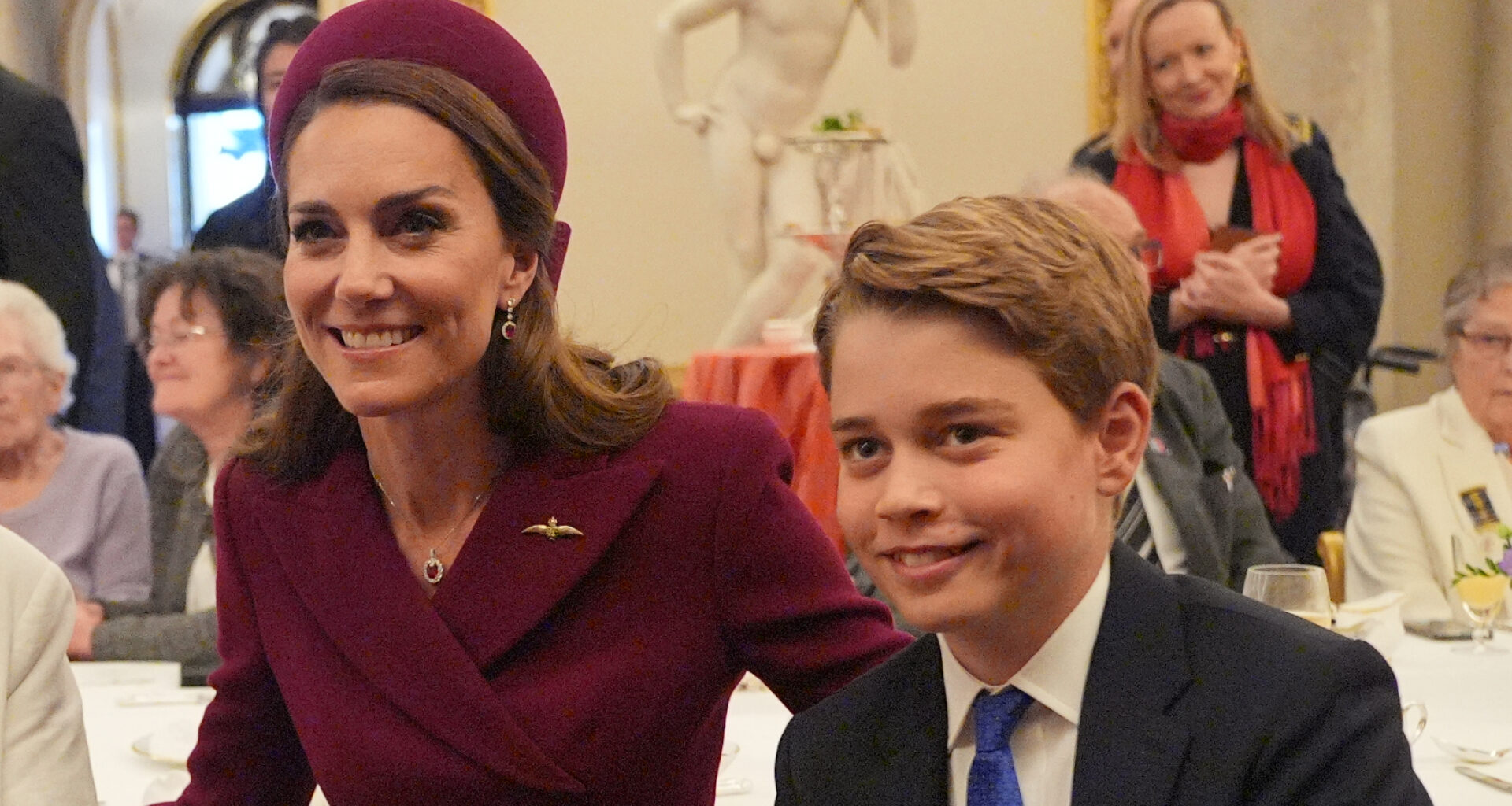 Kate Middleton wears a burgundy coat and matching hat and poses with son Prince George, who is wearing a navy suit, white shirt, and blue tie