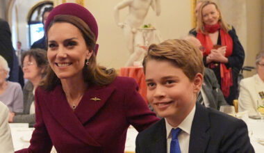 Kate Middleton wears a burgundy coat and matching hat and poses with son Prince George, who is wearing a navy suit, white shirt, and blue tie