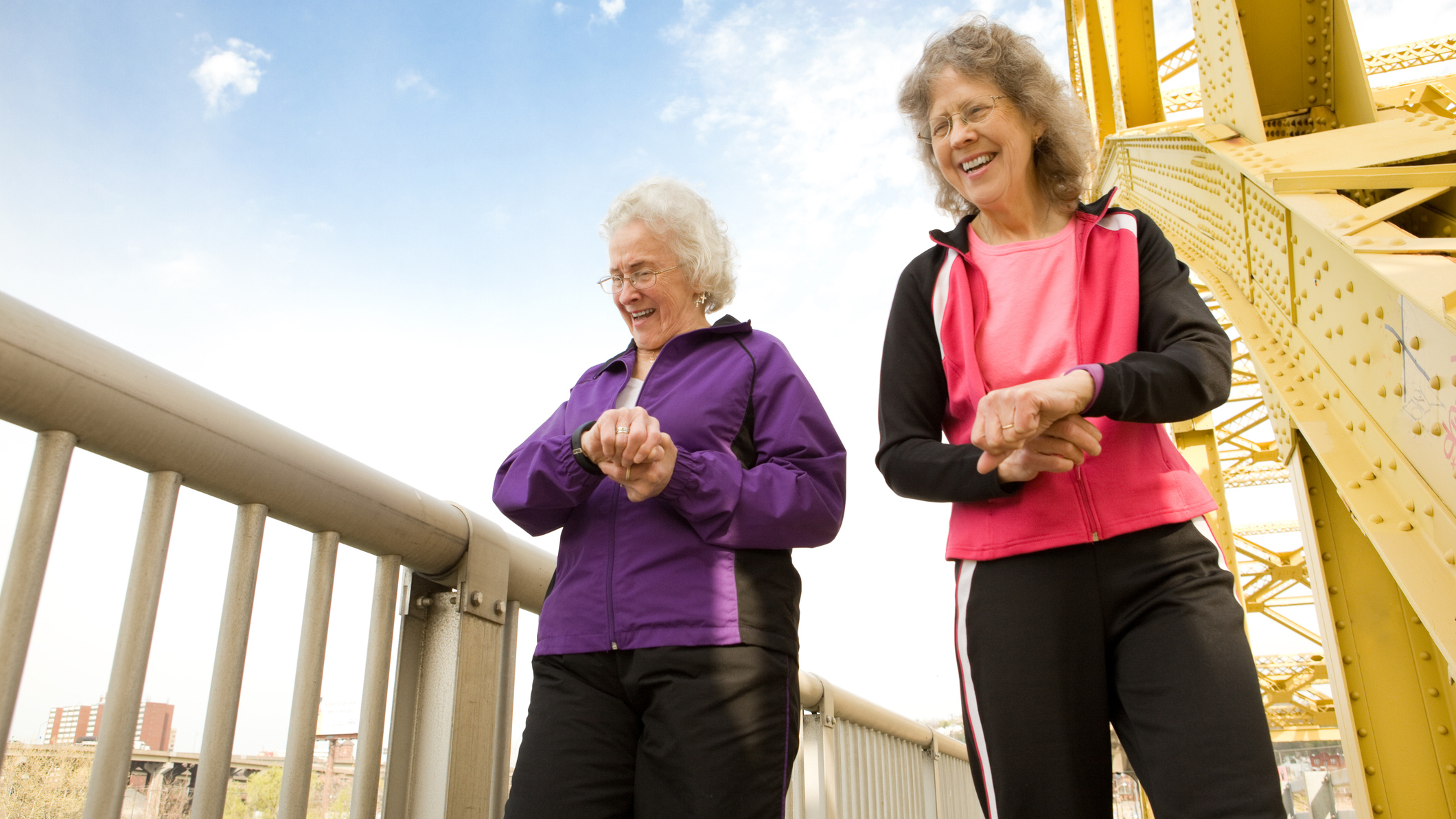 Two senior women laughing together as they check their step counters on the walkway of a bridge