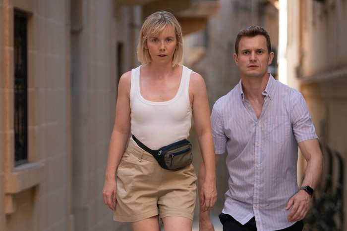 Lauren Lyle and Chris Jenks wearing light summer clothes, walking up stone steps with serious expressions on their faces as they walk through a narrow alleyway.