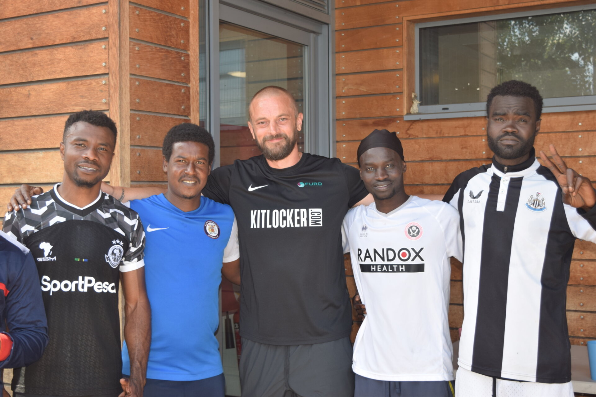 Five men stand in from of a wood panelled community centre building. They are all from different backgrounds and ethnicities. They look comfortable, content and friendly with each other.