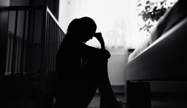 A woman sits in shadow next to a crib.