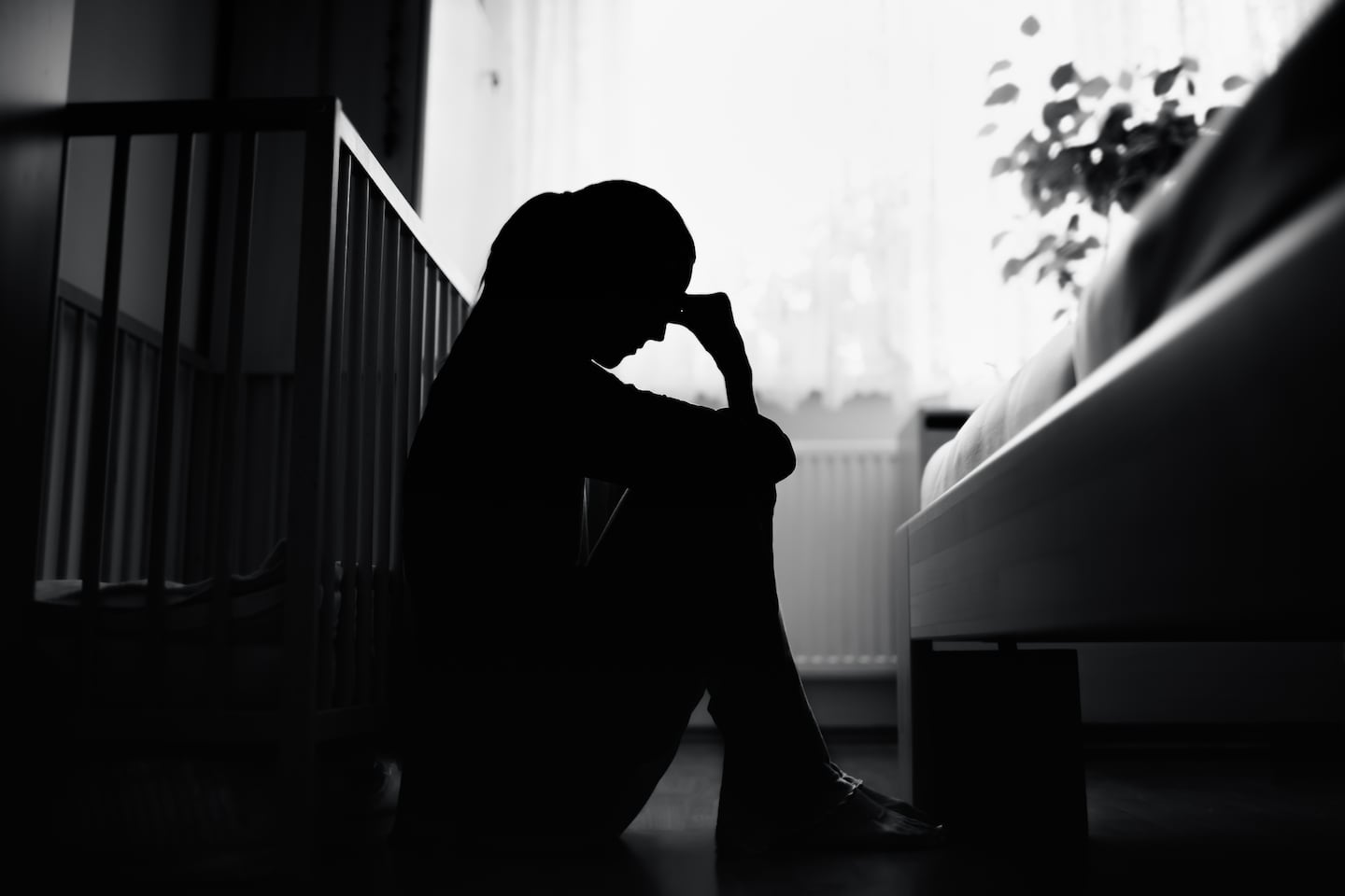 A woman sits in shadow next to a crib.