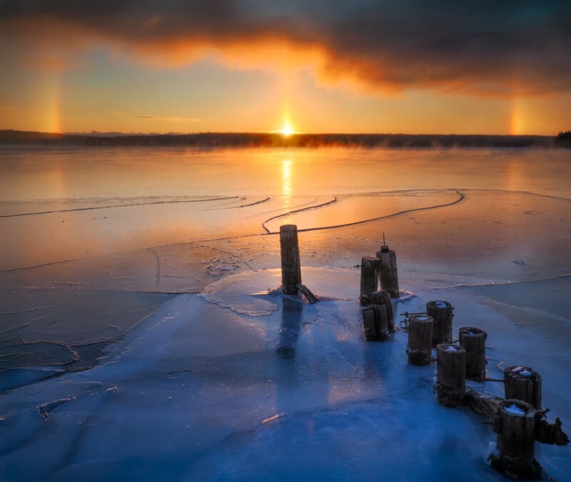 A frozen lake at sunrise with colorful sun dogs and steaming mist above the ice. Old wooden posts jut out of the icy surface in the foreground, reflecting the warm orange and blue hues of the sky.