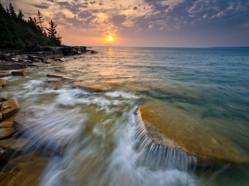 Sunset over a rocky shoreline with water flowing over flat stones into a clear lake, trees lining the left edge, and soft clouds glowing in the orange and purple sky.