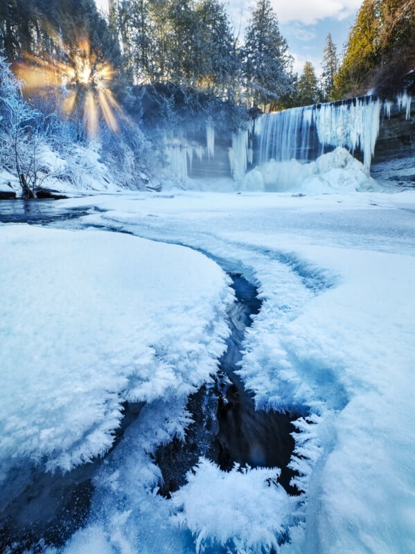 A frozen waterfall surrounded by snow-covered trees with sunlight streaming through the branches; icy patterns frame a narrow stream flowing beneath the frozen surface in the foreground.
