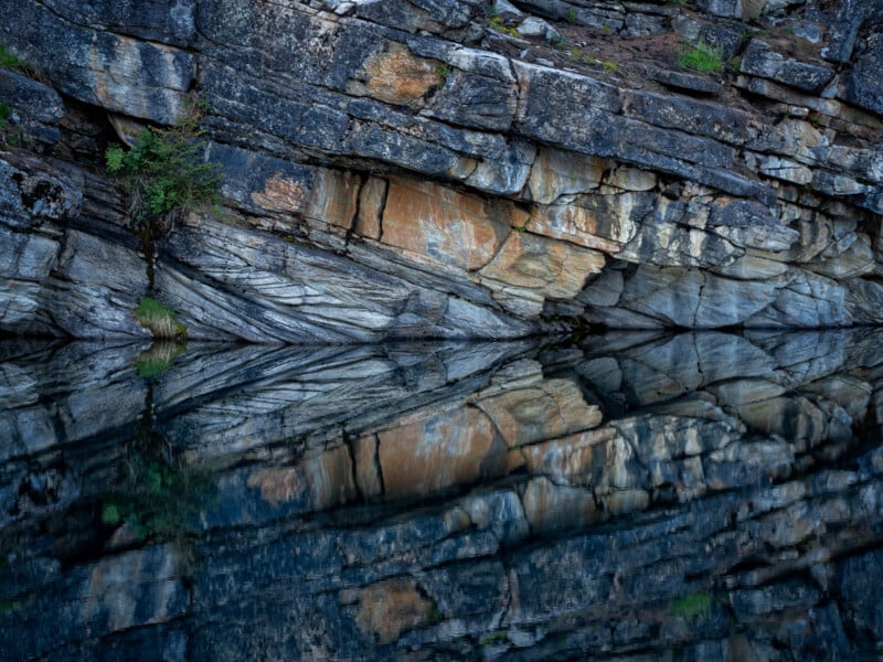 Jagged rock face with various shades of brown, gray, and orange, reflected clearly in the still water below; a few small green plants grow in cracks along the rock surface.