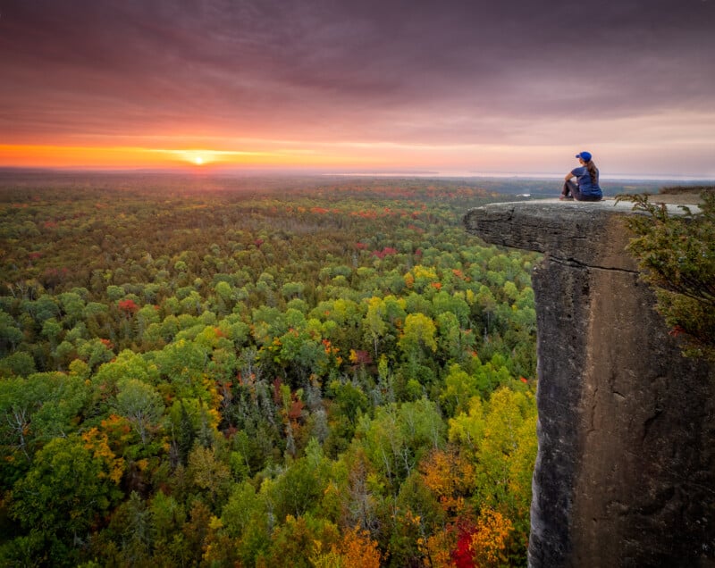 A person sits on the edge of a high cliff, overlooking a vast forest with colorful autumn foliage at sunrise. The sky is cloudy, with the sun casting an orange glow over the landscape.