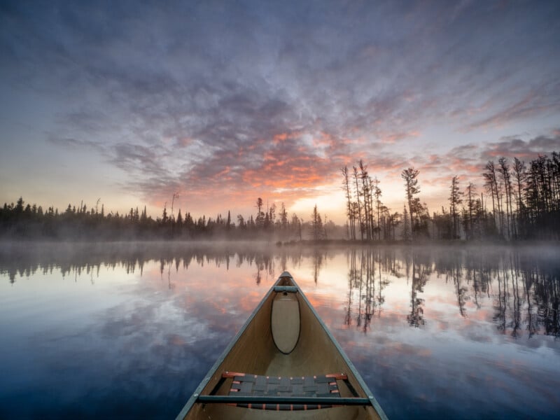 A canoe glides on a calm lake at sunrise, with mist rising from the water and silhouettes of trees reflected on the surface under a colorful, cloudy sky.