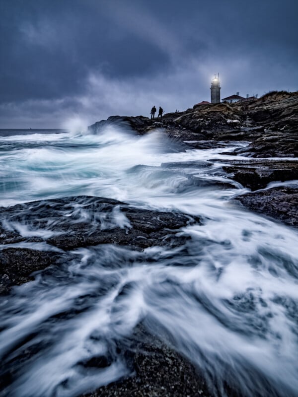 Waves crash onto rocky shore under a stormy sky, with two silhouetted figures near a lighthouse glowing on the distant cliff.