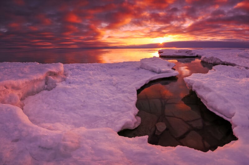 Sunset over icy shoreline with snow-covered rocks and patches of open water reflecting the vivid orange, pink, and purple colors of the dramatic sky.