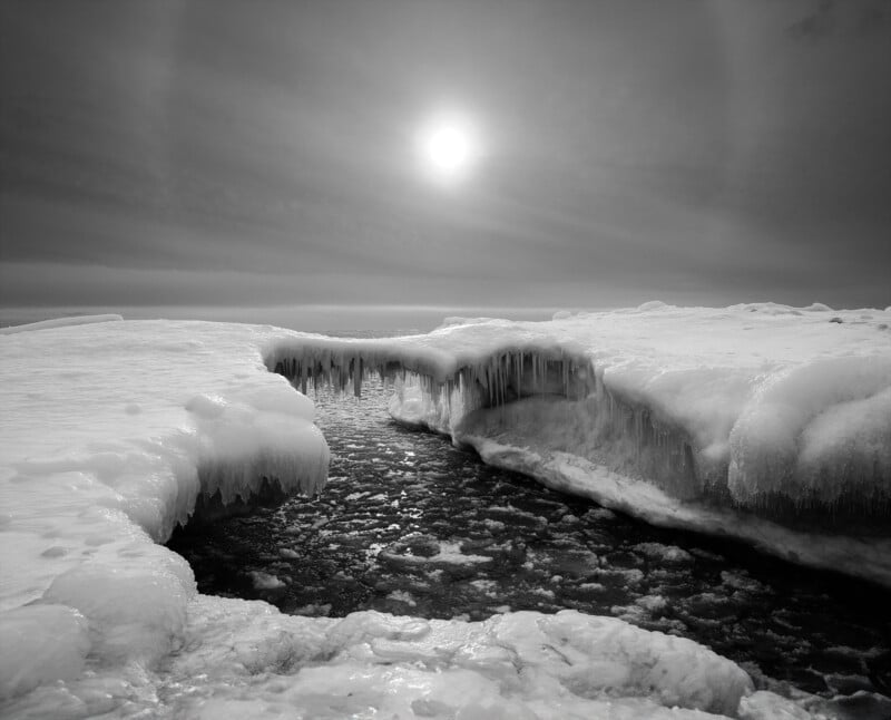 The black and white photo shows a frozen landscape with ice and snow surrounding a narrow, icy stream. The sun shines brightly in the cloudy sky, creating a dramatic, cold, and serene scene.