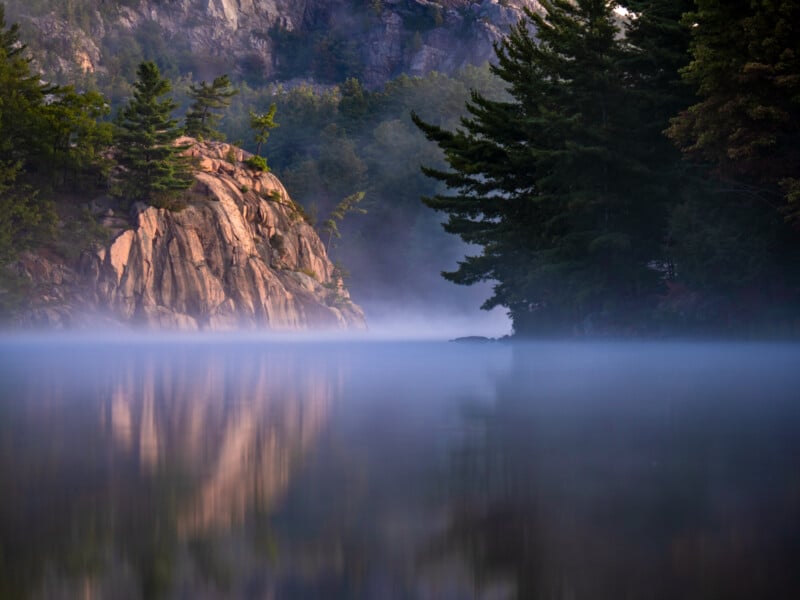 Fog rises over a calm lake surrounded by rocky cliffs and dense pine trees, with reflections of the landscape mirrored on the water’s surface and soft morning light illuminating the scene.