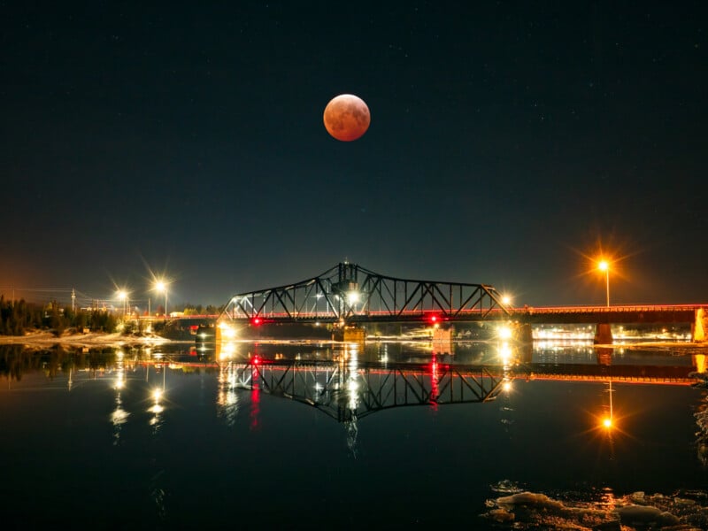 A large red moon hangs above a lit bridge at night, with streetlights shining and the bridge reflected in the calm river below, under a clear, starry sky.