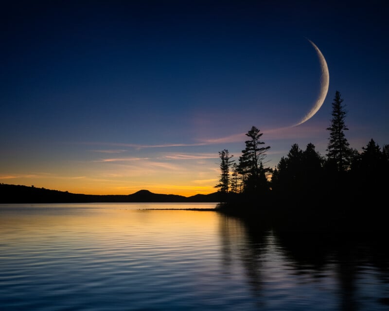 A crescent moon rises above a calm lake at sunset, with silhouettes of tall pine trees and distant hills reflected in the tranquil water under a vibrant, colorful sky.