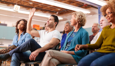Man putting his hand up in a meeting surrounded by a group of other people