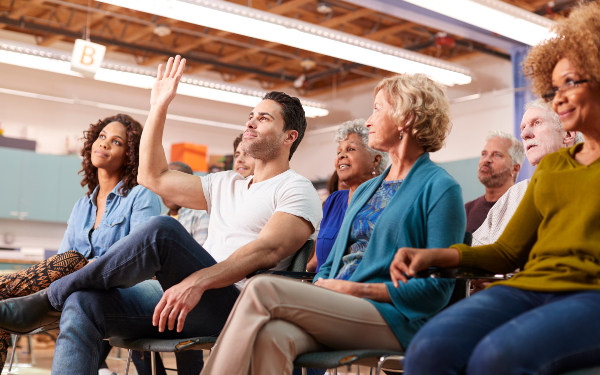 Man putting his hand up in a meeting surrounded by a group of other people