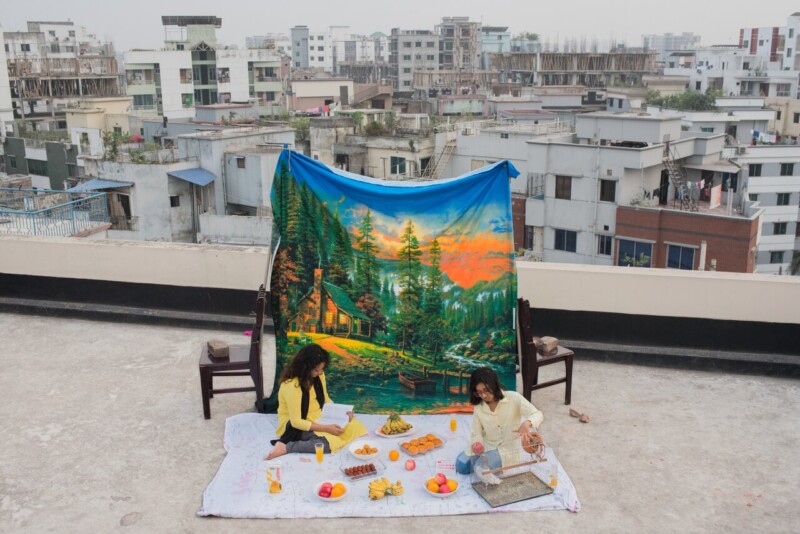Two people have a picnic on a rooftop, sitting on a blanket with food and books. Behind them is a large tapestry with a colorful nature scene, and city buildings are visible in the background.