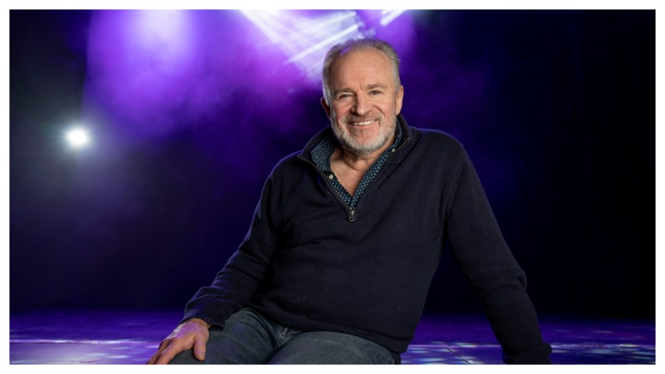 Smiling man sitting on stage under purple lighting.